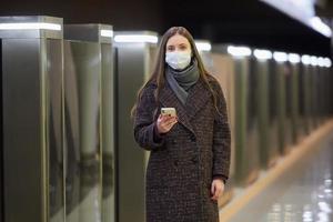 una mujer con una mascarilla médica está esperando un tren y sosteniendo un teléfono inteligente foto