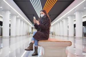 A woman in a medical face mask is waiting for a train and holding a smartphone photo