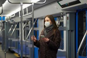 A woman in a face mask is standing and using a smartphone in a modern subway car photo