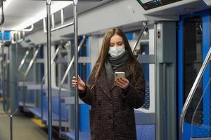 A woman in a face mask is standing and using a smartphone in a modern subway car photo