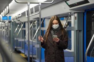 una mujer con una mascarilla está de pie y usando un teléfono inteligente en un vagón de metro moderno foto