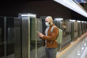 un hombre con una máscara facial está usando un teléfono inteligente mientras espera un tren subterráneo foto