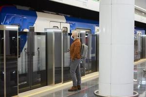 A man in a face mask is holding a smartphone while waiting for a subway train photo