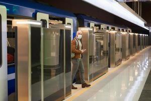 El hombre con una mascarilla médica sostiene un teléfono mientras deja un vagón de metro moderno foto