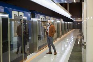 A man in a face mask is holding a smartphone while waiting for a subway train photo