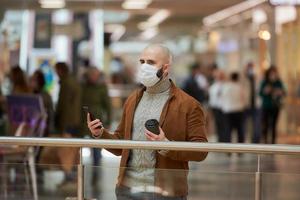 El hombre con una mascarilla está usando un teléfono y sosteniendo un café en el centro comercial. foto