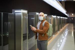 A man in a face mask is using a smartphone while waiting for a subway train photo