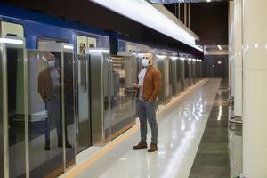 un hombre con una máscara facial sostiene un teléfono inteligente mientras espera un tren subterráneo foto