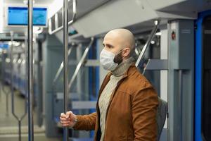 Un hombre calvo con barba en una mascarilla está sosteniendo el pasamanos en un vagón de metro foto