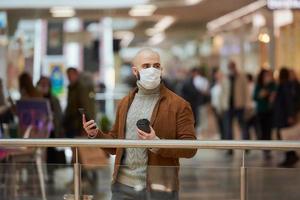 El hombre con una mascarilla está usando un teléfono y sosteniendo un café en el centro comercial. foto