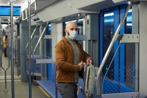 A bald man with a beard in a face mask is putting on a backpack in a subway car photo