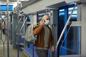A bald man with a beard in a face mask is putting on a backpack in a subway car photo
