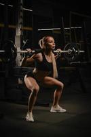 una mujer en forma está en cuclillas con una barra cerca de la rejilla de sentadillas en un gimnasio
