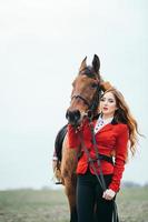 Red-haired jockey girl in a red cardigan and black high boots with a horse photo