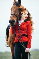 Red-haired jockey girl in a red cardigan and black high boots with a horse photo