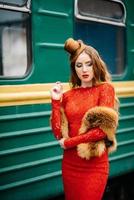 Chica joven con cabello rojo con un vestido rojo brillante cerca de un viejo automóvil de pasajeros foto