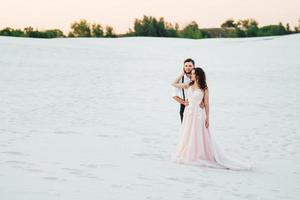 young couple a guy in black breeches and a girl in a pink dress are walking along the white sand photo