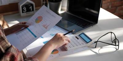 Close up of businessman or accountant hand holding pen working on calculator to calculate business data, accountancy document and laptop computer at office, business concept photo