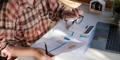 Cerca de la mano de la empresaria o del contador que sostiene la pluma trabajando en la calculadora para calcular los datos comerciales, el documento de contabilidad y la computadora portátil en la oficina, concepto de negocio foto