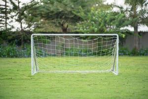 El enfoque selectivo en una pequeña portería de fútbol con red de cuerda pone en el campo de hierba con jardín borrosa y árboles en segundo plano. foto