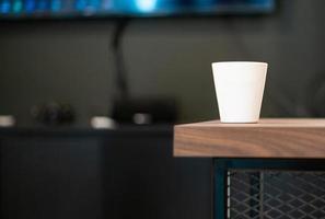 Closeup white cup without screen on the surface for adding some text puts on the wooden table with blurred electronic equipments in background photo