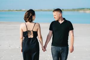 Young couple a guy with a girl in black clothes are walking on the white sand photo