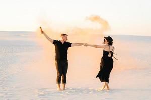 Guy and a girl in black clothes hug and run on the white sand with orange smoke photo