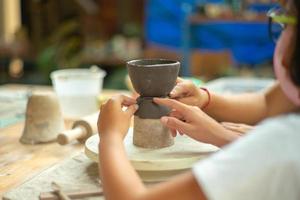 Closeup hands of the teacher and student helping to mold the clay jar in a workshop classroom with blurred people in the background. photo