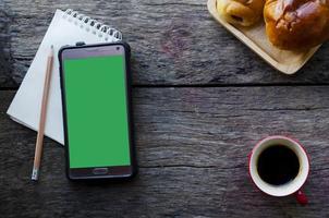 Smartphone con pantalla verde y bloc de notas con lápiz y taza de café roja sobre fondo de madera foto