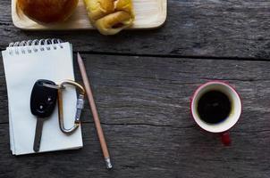 Car keys on notepad with pencil and red coffee cup and bun bread on wood table background photo
