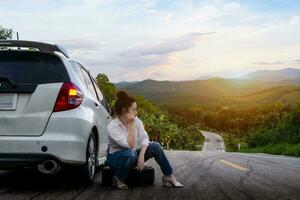 Young beautiful lady sitting near a car for calling for help on the public road in a forest area with a mountain and sky background photo