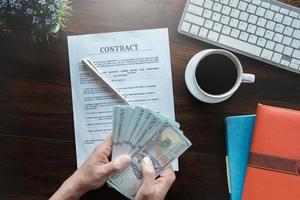 concepto de contrato y acuerdo, hombre que sostiene la banda de dólar y firma el papel de solicitud de contrato con el teclado de la taza de café y el cuaderno en el escritorio de madera en el banco de la oficina. foto