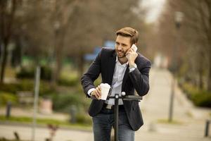 empresario casual montando un scooter mientras sostiene un café y habla por teléfono