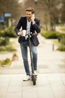 empresario casual sosteniendo un café y hablando por teléfono mientras está de pie en un scooter
