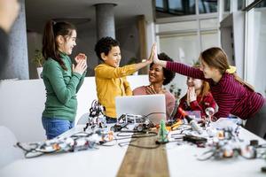 Niños felices con su profesora de ciencias afroamericana con programación de portátiles, juguetes eléctricos y robots en el aula de robótica foto