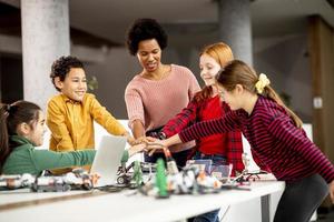 Niños felices con su profesora de ciencias afroamericana con programación de portátiles, juguetes eléctricos y robots en el aula de robótica