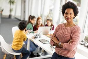 Profesora de ciencias afroamericana con un grupo de niños que programan juguetes eléctricos y robots en el aula de robótica foto