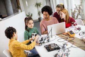 Niños felices con su profesora de ciencias afroamericana con programación de portátiles, juguetes eléctricos y robots en el aula de robótica foto