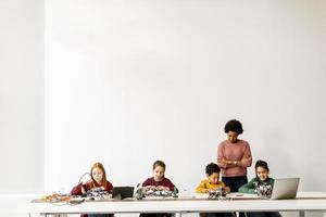 Niños felices con su profesora de ciencias afroamericana con programación de portátiles, juguetes eléctricos y robots en el aula de robótica foto