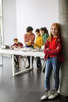 Cute little girl standing in front of kids programming electric toys and robots at robotics classroom photo