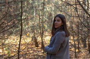 Chica medio vuelta con un abrigo gris que se encuentra en el bosque en un soleado día de otoño