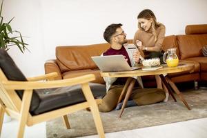 Young woman and young man using laptop for onlin payment while sitting by sofa at home