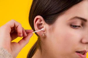 Young woman cleaning her ears with cotton sticks on yellow background photo