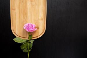 Una rosa rosa descansa sobre una tabla de cortar de madera sobre un fondo oscuro