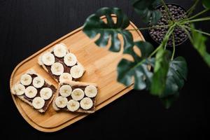 Three banana white bread toasts spread with chocolate butter on a chopping board with leaves on a dark background photo