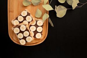 Three banana white bread toasts spread with chocolate butter on a chopping board with leaves on a dark background photo