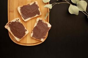 Three banana white bread toasts spread with chocolate butter on a chopping board with leaves on a dark background photo