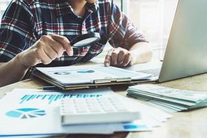 Business man using magnifying to review balance sheet annual with using laptop computer to calculating budget photo