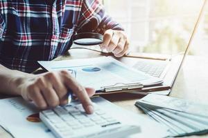 Business man using magnifying to review balance sheet annual with using calculator and laptop computer to calculating budget photo