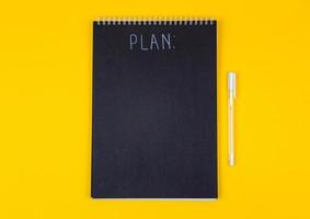 Blank black spiral note pad and white marker with calligraphic inscription of the word plan on yellow background